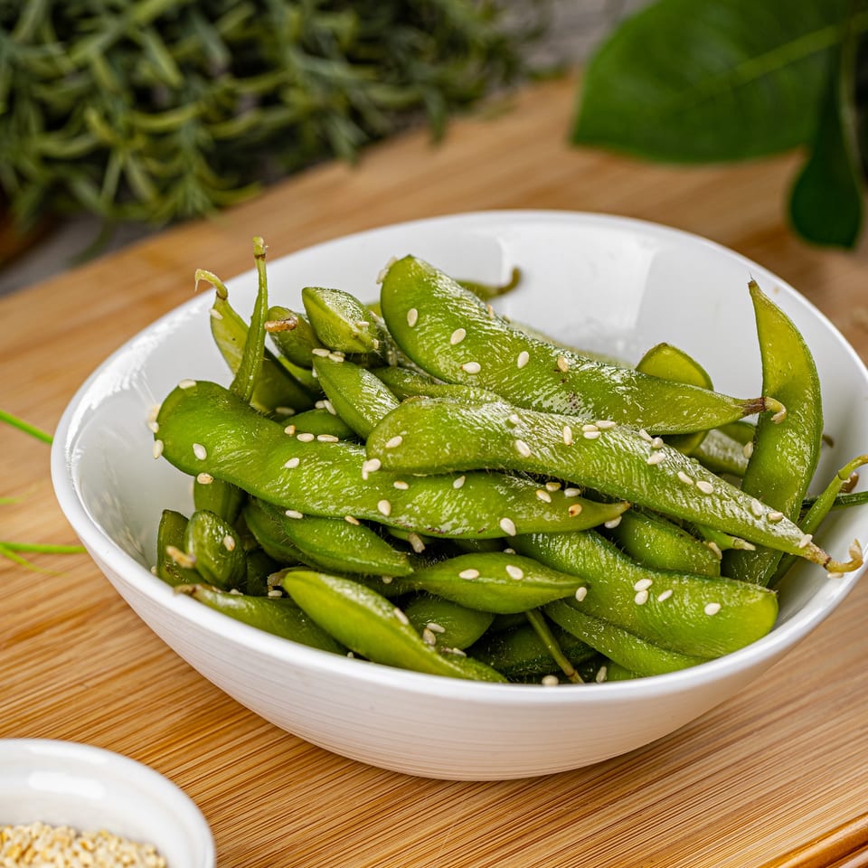 A white bowl filled with vibrant green edamame pods, sprinkled with white sesame seeds, on a wooden surface.