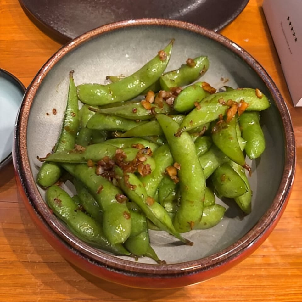 A bowl of vibrant green edamame pods, sprinkled with red chili flakes, on a wooden table.
