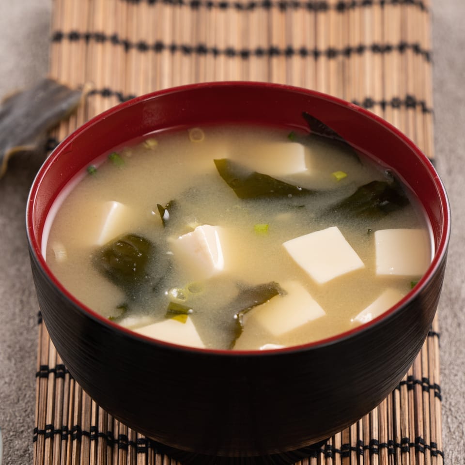A black and red bowl of Japon miso soup with tofu and seaweed on a bamboo mat.
