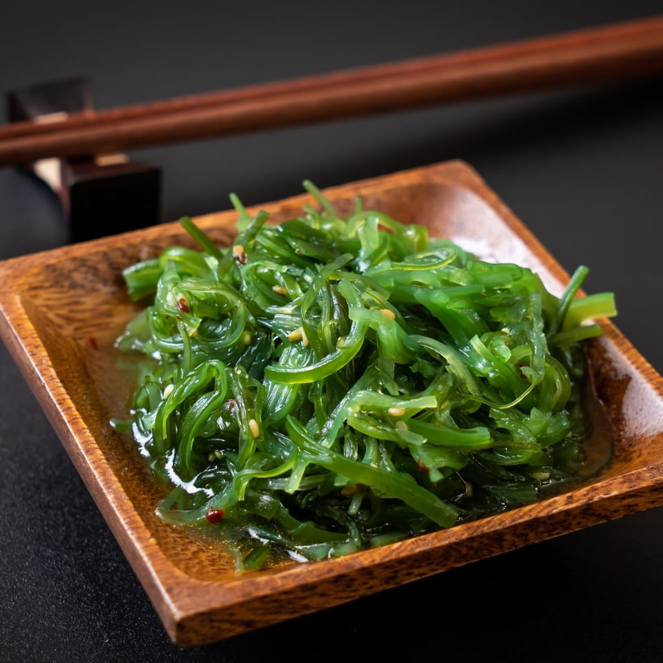 Bright green seaweed salad with sesame seeds in a wooden bowl, with chopsticks in the background.