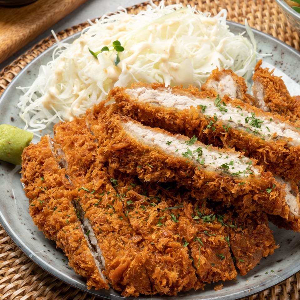 Crispy, sliced Japanese tonkatsu with shredded cabbage salad and a dollop of wasabi on a grey plate.
