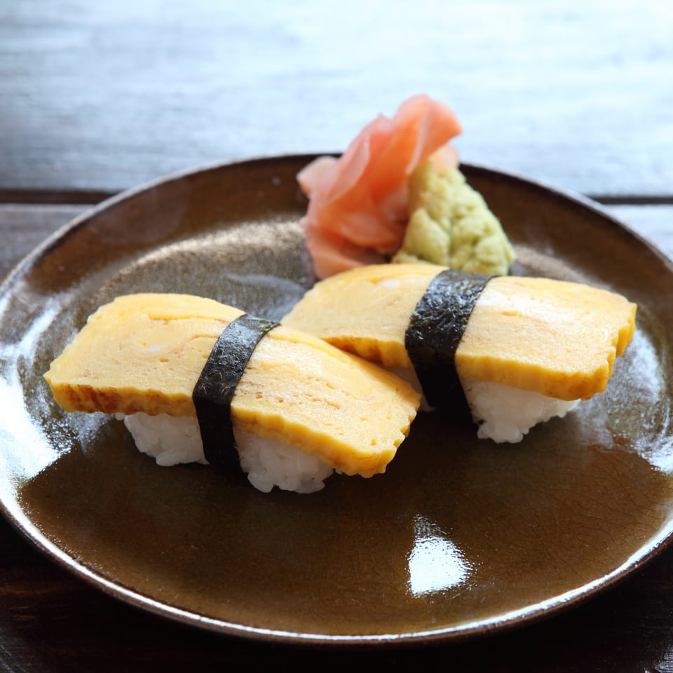 Two pieces of tamago nigiri sushi with pickled ginger and wasabi on a brown plate.